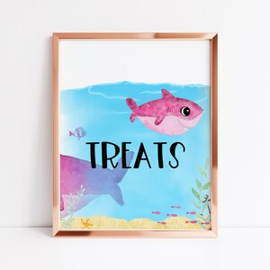 May include: A framed watercolor print featuring a pink cartoon shark swimming in blue water. The word "TREATS" is written in black. The background includes a sand-colored seabed, fish, and aquatic plants.