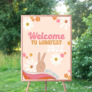 Peut inclure: Une pancarte d'accueil rose et orange avec un signe de paix et des fleurs. La pancarte indique "Bienvenue &agrave; LunaFest 2022" et "Let's Party !"