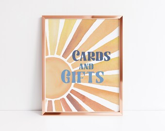 May include: A framed print with a watercolor sunburst design in shades of orange, yellow, and brown. The text "CARDS AND GIFTS" is written in blue letters on the sun.