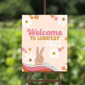 Peut inclure: Une pancarte d'accueil rose et blanche avec un signe de paix et des fleurs. La pancarte indique "Bienvenue &agrave; LunaFest 2022" et "Let's party !"