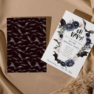 May include: A white baby shower invitation with a black coffin-shaped frame decorated with blue and pink flowers and bats. The text on the invitation reads "Oh Baby!" and includes the name "Miranda Jones".