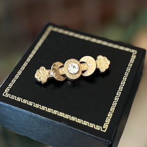 May include: A gold-toned brooch with a central clear gemstone and decorative circular and floral elements. The brooch is set on a black box with a gold border, suggesting a jewelry presentation.