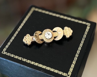 May include: A gold-toned brooch with a central clear gemstone and decorative circular and floral elements. The brooch is set on a black box with a gold border, suggesting a jewelry presentation.
