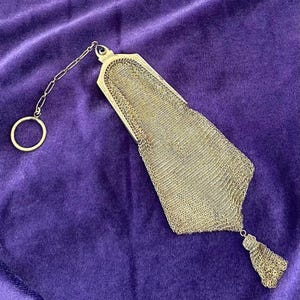 May include: A vintage silver mesh purse with a chain handle and a tassel. The purse is rectangular with a decorative top and a delicate chain link design. The background is a deep purple velvet.
