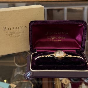 May include: A gold-tone Bulova Fifth Avenue wristwatch with a delicate link bracelet, presented in a dark purple velvet-lined display case. The box lid features the text Bulova Fifth Avenue New York, matching the branding inside the case.