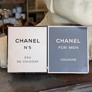 May include: A rectangular Chanel N°5 Eau de Cologne and Chanel For Men Cologne product label. The left side is cream with black text, and the right side is grey with white text. The two sections are joined by a small white metal fastener.