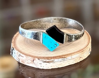 May include: A silver bangle bracelet with a turquoise and black stone design. The bracelet is on a wooden surface.