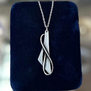 May include: A silver-toned pendant necklace with an abstract design. The pendant features a geometric shape and an infinity symbol, suspended from a delicate chain. The necklace is displayed on a dark blue velvet surface.