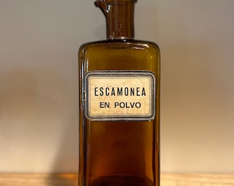 May include: A brown glass bottle with a label that reads "ESCAMONEA EN POLVO".