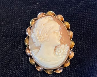 Puede incluir: Broche de camafeo ovalado con el perfil de una mujer tallado en blanco sobre un fondo melocotón. El broche está enmarcado por un borde festoneado en tono dorado. Una joya clásica.