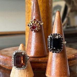 May include: Three vintage rings displayed on wooden stands. The rings feature large gemstones in red, black, and brown, set in gold and silver-toned metal. The rings are showcased on small, wooden, cone-shaped displays.