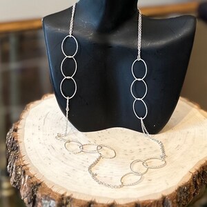 May include: A silver chain necklace with multiple interconnected oval links. The necklace is displayed on a wooden stand.