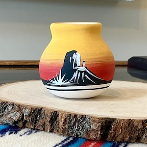 Puede incluir: Un pequeño jarrón de cerámica pintado a mano con un diseño de paisaje. El jarrón presenta una parte superior amarilla, que transiciona a rojo y naranja, con una silueta de montaña negra y detalles blancos. La base es blanca.