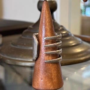 May include: A silver ring with a rectangular face and multiple curved bands is displayed on a wooden ring holder. The ring holder is a cone shape and is made of a reddish-brown wood. The background includes a brass lamp.