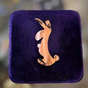 Op de afbeelding: Een koperkleurige konijnenhanger met een lachend gezicht, tegen een donkerpaarse fluwelen achtergrond. Het konijn is gestileerd met een gebogen lichaam en lange oren. De hanger is waarschijnlijk een sieraad.