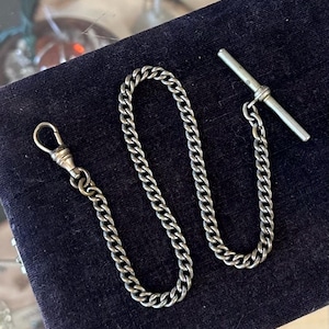 May include: A silver-toned pocket watch chain with a T-bar and a clasp. The chain is made of interlocking oval links. The chain is displayed on a dark blue velvet surface.