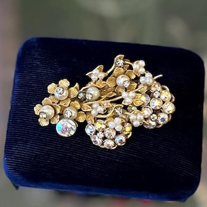 Puede incluir: Un broche dorado con un diseño floral que presenta perlas blancas y diamantes de imitación transparentes. El broche se muestra sobre un fondo de terciopelo azul.