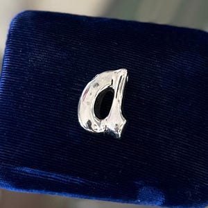 May include: A silver letter "A" charm, possibly a pendant or brooch, set against a dark blue velvet backdrop. The letter has a textured surface, reflecting light. The item is likely jewellery, with a focus on the initial.