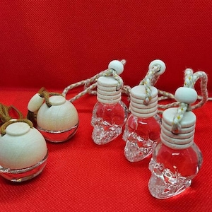 Könnte beinhalten: Vier Auto-Lufterfrischer, die an Schnüren hängen. Drei sind schädelförmige Glasflaschen mit weißen Holzverschlüssen. Eine ist eine runde Glasflasche mit einem weißen Holzverschluss.