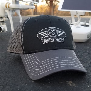 May include: A black and gray mesh trucker hat with a white embroidered patch that says "FAA UAS 107 DRONE PILOT".