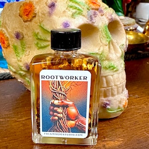 May include: A clear glass bottle filled with a golden liquid and herbs. The bottle has a black label with the text "ROOTWORKER" and a graphic of a hand holding a bundle of roots. The label also includes the website "folkmagicstudio.com".