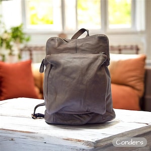 Puede incluir: Una mochila de cuero marrón con un bolsillo frontal con cremallera y un asa superior. La bolsa tiene correas ajustables y un cierre de solapa. La mochila está sobre una superficie de madera blanca. La palabra "Conders" está en la esquina inferior derecha.