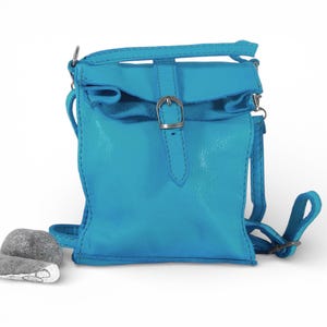 Puede incluir: Un bolso bandolera de cuero en color turquesa brillante con un cierre de solapa plegable asegurado mediante una hebilla de metal plateado. El bolso incluye una correa ajustable a juego sujeta con herrajes plateados y costuras visibles.