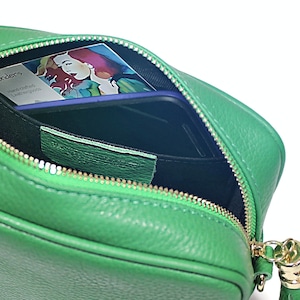 May include: A green leather crossbody bag with a gold zipper and tassel. The bag is open and shows a black interior with a small card and a phone inside.