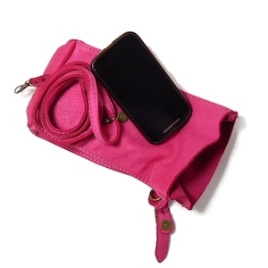Puede incluir: Un bolso bandolera de cuero rosa brillante con superficie texturizada, que cuenta con una correa enrollada a juego y un cierre de presi&oacute;n de metal en tono lat&oacute;n. Un smartphone negro descansa sobre el bolso, que tiene una abertura superior y forma rectangular.