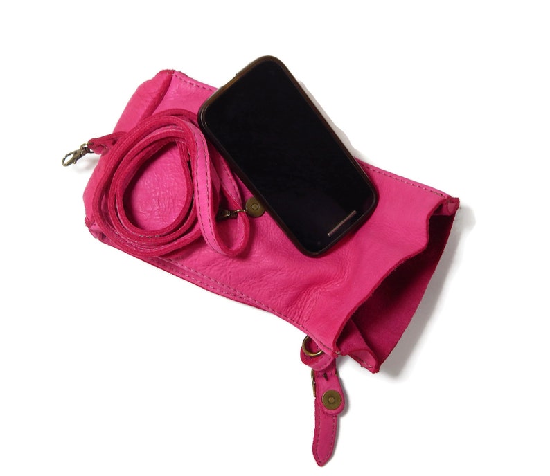Puede incluir: Un bolso bandolera de cuero rosa brillante con superficie texturizada, que cuenta con una correa enrollada a juego y un cierre de presi&oacute;n de metal en tono lat&oacute;n. Un smartphone negro descansa sobre el bolso, que tiene una abertura superior y forma rectangular.