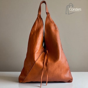 May include: A large, orange-brown leather tote bag with a unique, asymmetrical design. The bag has a single handle and a drawstring closure. The bag is made of a soft, supple leather and has a minimalist aesthetic.