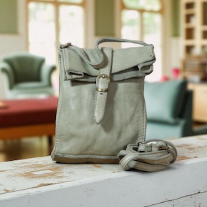 Puede incluir: Un bolso bandolera de cuero color verde salvia con una solapa superior plegable asegurada por una correa vertical y una hebilla de metal color lat&oacute;n. El bolso tiene una forma suave y desestructurada e incluye una correa fina de cuero a juego.