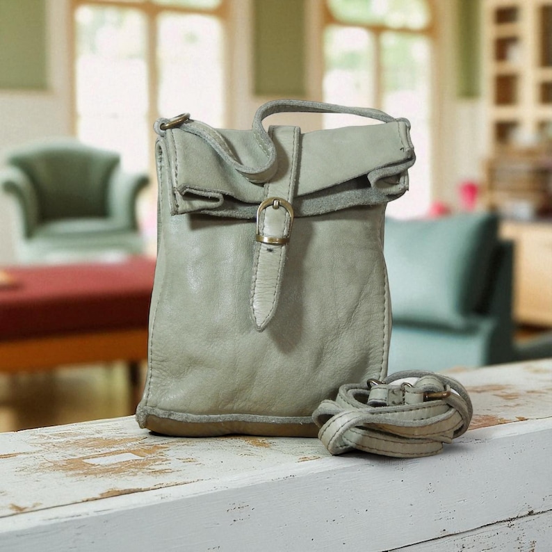 Puede incluir: Un bolso bandolera de cuero color verde salvia con una solapa superior plegable asegurada por una correa vertical y una hebilla de metal color lat&oacute;n. El bolso tiene una forma suave y desestructurada e incluye una correa fina de cuero a juego.