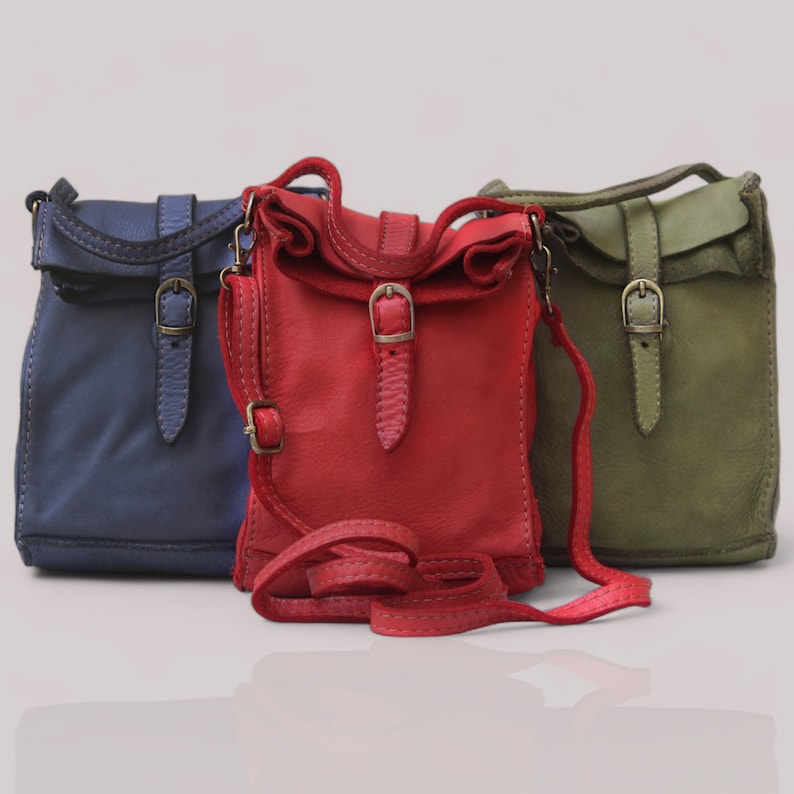 Puede incluir: Tres bolsos bandolera peque&ntilde;os en azul, rojo y verde oliva. Cada bolso cuenta con una solapa superior, una correa vertical con hebilla de metal en tono lat&oacute;n y una correa de hombro ajustable a juego con un peque&ntilde;o deslizador de metal.