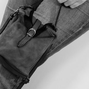 Puede incluir: Un bolso bandolera peque&ntilde;o de cuero en color oscuro, con forma rectangular y superficie texturizada. El bolso presenta una correa vertical central asegurada por una hebilla de metal plateado, correas laterales finas y una correa larga.