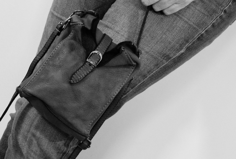 Puede incluir: Un bolso bandolera peque&ntilde;o de cuero en color oscuro, con forma rectangular y superficie texturizada. El bolso presenta una correa vertical central asegurada por una hebilla de metal plateado, correas laterales finas y una correa larga.