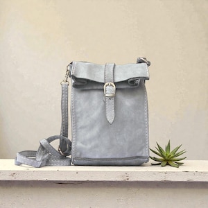 Puede incluir: Un bolso bandolera de color gris claro con cierre enrollable asegurado por una correa vertical y una hebilla de metal plateado. El bolso cuenta con una correa larga y fina a juego, sujeta a los lados con herrajes de metal plateado.
