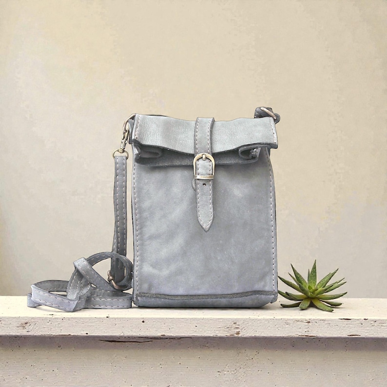 Puede incluir: Un bolso bandolera de color gris claro con cierre enrollable asegurado por una correa vertical y una hebilla de metal plateado. El bolso cuenta con una correa larga y fina a juego, sujeta a los lados con herrajes de metal plateado.