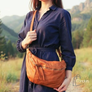 Puede incluir: Un bolso bandolera de cuero marrón con forma curva y cremallera dorada. El bolso tiene una correa larga y se lleva al hombro. El bolso es de color marrón cálido y parece estar hecho de cuero suave. Un accesorio elegante.