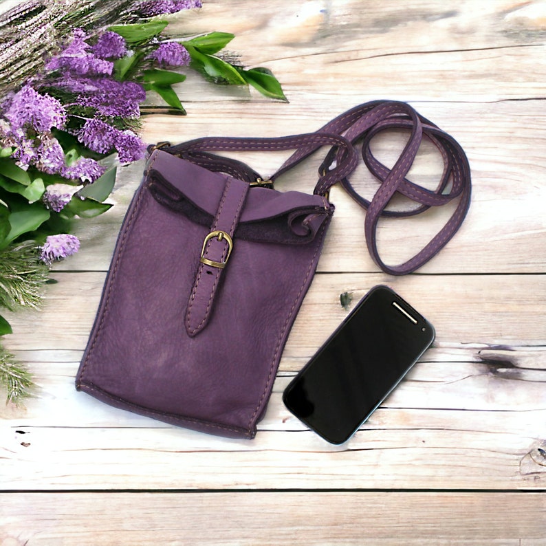 Puede incluir: Un bolso bandolera de cuero morado con solapa plegable, cierre de hebilla en tono lat&oacute;n y una correa larga a juego. El bolso presenta costuras visibles en los bordes y est&aacute; colocado junto a un tel&eacute;fono inteligente negro con pantalla apagada.