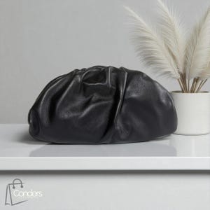 May include: A black leather clutch bag with a gathered design. The bag is oval-shaped and sits on a white surface. A white vase with decorative plumes is in the background.
