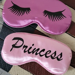 May include: Two pink satin sleep masks. One mask has black eyelashes printed on it. The other mask has the word "Princess" printed on it in black.