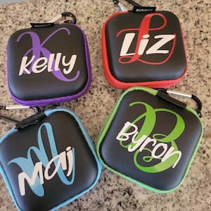 May include: Four black zippered pouches with a carabiner clip. Each pouch has a different name written in a decorative font on the front. The names are Kelly, Liz, Maj, and Byron. The pouches are in different colors: purple, red, blue, and green.