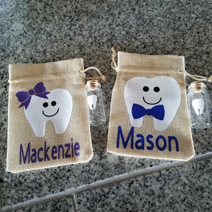 May include: Two tooth-shaped burlap bags with personalized names, Mackenzie and Mason, each with a small glass vial containing a tooth. The bags have a white tooth with a smiling face, a bow, and a bow tie.