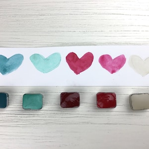 Puede incluir: Cinco pastillas de pintura acuarela con muestras de pintura verde azulado, turquesa, rojo, rosa y blanco. Las muestras de pintura tienen forma de corazón.