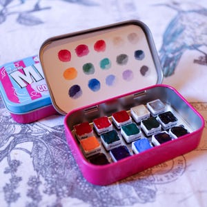 Puede incluir: Una lata de metal rosa con un juego de 15 acuarelas. La lata tiene una tapa de plástico transparente con un inserto de papel blanco que muestra los colores de las pinturas. Los colores de la pintura incluyen rojo, escarlata, titanio, azul oscuro, gris raro, verde esmeralda, azul de Prusia, y más.