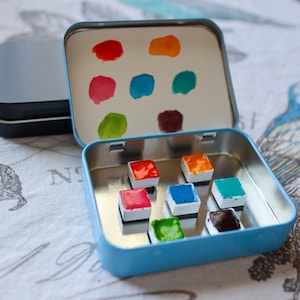 Puede incluir: Una lata de metal azul con una tapa que se abre para revelar un juego de nueve pastillas de pintura de acuarela. Las pastillas están dispuestas en una cuadrícula de 3x3 y están llenas de diferentes colores, incluyendo rojo, naranja, amarillo, verde, azul y morado. La tapa de la lata tiene una superficie de espejo y muestra los colores de las pastillas de pintura.