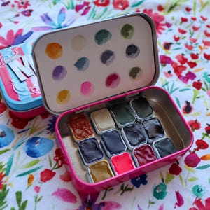 Puede incluir: Una lata de metal rosa con una tapa que se abre para revelar un juego de 12 pastillas de pintura de acuarela. La tapa tiene un fondo blanco con un patrón de círculos coloridos. Las pastillas de pintura están dispuestas en una cuadrícula y están llenas de diferentes colores de pintura.