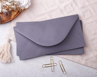 May include: Three grey envelopes with curved flaps, arranged on a textured, off-white surface. Two gold paper clips are placed below. A wooden tray with dried flowers and a tassel are also visible.