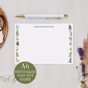 May include: A6 personalised stationery with a fern leaf border. The white paper has the name "HARPER REDSHAW" printed at the top. A white pen with gold accents rests above the stationery. A green circle with the text "PERSONALISE ADD ANY NAME" is in the lower left corner.